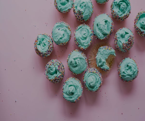 A group of cupcakes with light blue frosting and multicolored sprinkles is arranged on a light pink surface, ready for celebrations. One cupcake has a bite taken out of it, revealing the cake inside.