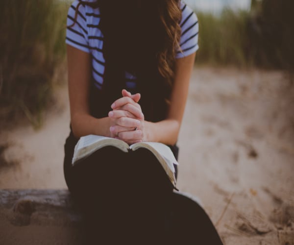 A person sits on a sandy beach, wearing a striped shirt and clasping their hands over an open book—a quiet celebration of solitude. The background is softly blurred, highlighting the blend of grass and sand.