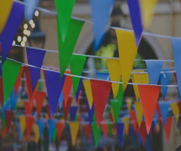 Colorful triangular pennant flags in blue, yellow, red, and green hang diagonally across an outdoor space. The background is blurred, suggesting a festive atmosphere with lights and a building partly visible—perfect for lively celebrations.