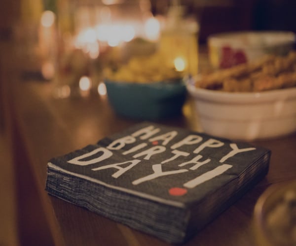 A stack of napkins with Happy Birthday! in bold sits on a table, setting the tone for joyful celebrations. In the background, blurred bowls of snacks and softly glowing candles add to the warm and festive atmosphere.