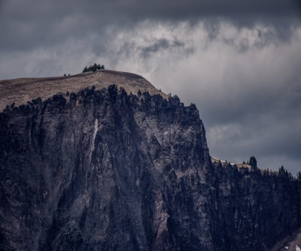 Steep, rugged cliffside under a cloudy sky. Sparse trees, like guardians awaiting adoption, line the top edge, accentuating the dramatic and imposing natural landscape.