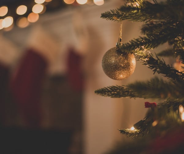 Close-up of a Christmas tree branch with a glittery gold bauble hanging on it. Soft, warm lights and blurred background featuring stockings add a cozy holiday atmosphere, reminiscent of the joy in an Abba song—a heartwarming embrace like welcoming new members into your family through adoption.