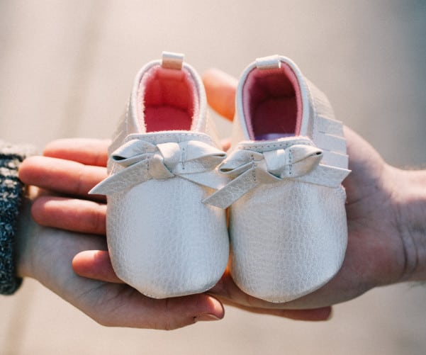 Two hands hold a pair of small white baby shoes with pink interiors and bow decorations, embodying the anticipation and joy found at an adoption agency. The softly blurred background highlights the shoes, symbolizing a new journey.