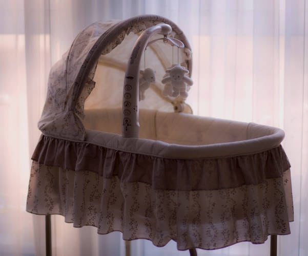 A baby bassinet with a sheer canopy sits near a sunlit window, symbolizing hope in the adoption process. It features a ruffled skirt and a mobile with two plush toys hanging from a curved arm. The soft lighting creates a serene atmosphere.