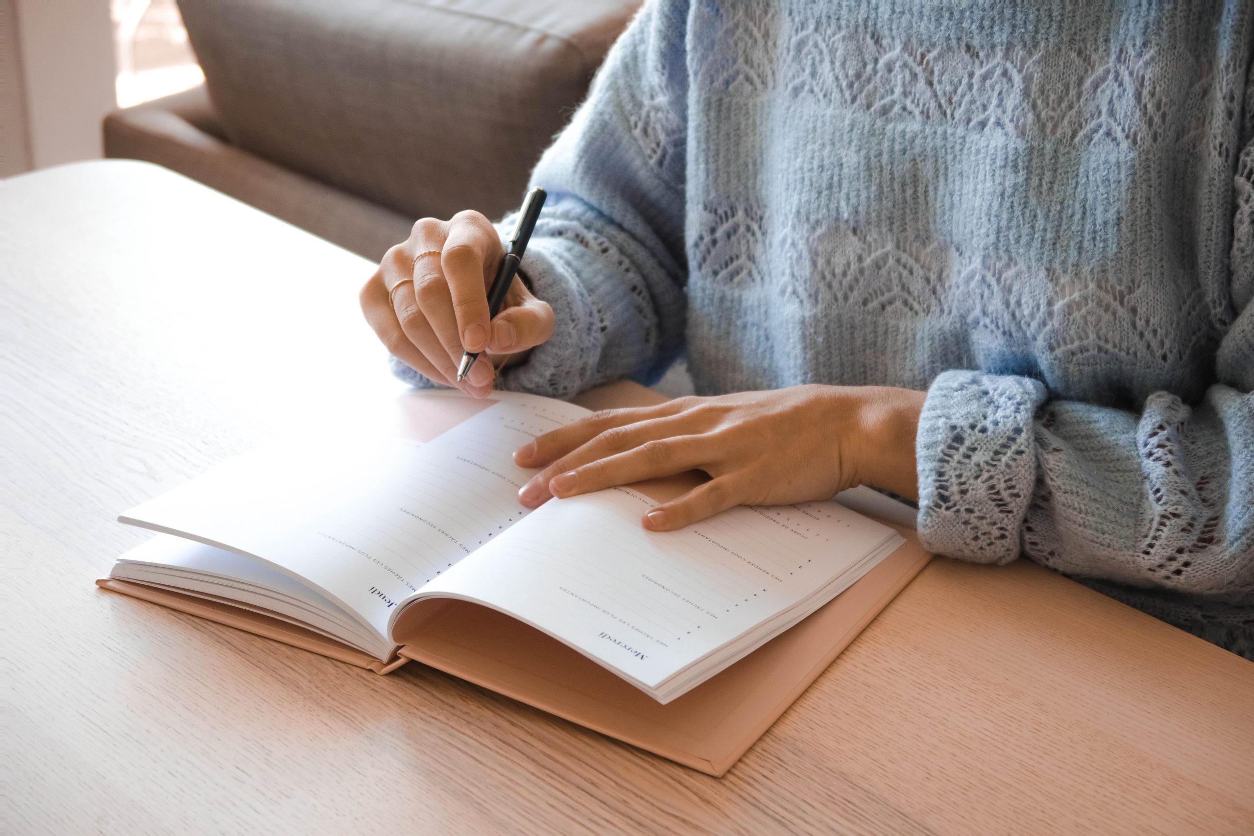 A person wearing a light blue sweater is sitting at a wooden table, writing in a notebook with a black pen. The notebook is open, showing blank pages. Furniture and soft lighting are in the background.